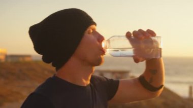 Sportif adam gün doğumunda denize bakan şişeden su içiyor ve gökyüzü berrak. Genç erkek koşucu şafakta mola verirken başka tarafa bakıyor.. 