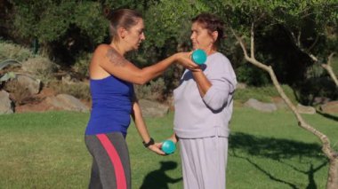 Kadın antrenör, yaşlı bir kadına parkta halterli egzersiz yaptırıyor. Antrenör rehberlik ve destek sunarken, idman kıyafeti giymiş yaşlı kadın egzersizlere odaklanıyor. 