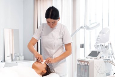 Güzellik kliniğinde kadın müşterileri temizleyen dermatolog. Rahatlamış genç bir kadın güzellik salonunda yüz bakımı görüyor.. 