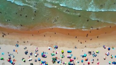 Ziyaretçiler parlak şemsiyeler altında kumlu sahilde dinlenirken, dalgalar kıyı boyunca çarpışıyor ve canlı bir yaz günü hava manzarasının özünü yakalıyor.