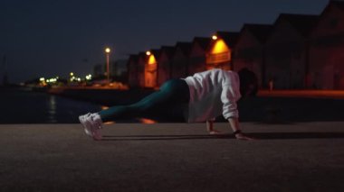 Karanlık şehirde geceleri sokaklarda şınav çeken kararlı, sportif bir kadın. Arka planda aydınlatılmış binalarla egzersiz yapan sağlıklı genç bir kadın vücudu. 