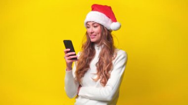 Mutlu Noel Baba kadın sarı arka planda dururken akıllı telefon kullanıyor. Güzel bir kadının portresi düz stüdyoda alet kullanıyor. 