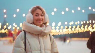 Kadın, Noel parkında parlak ışıklarla süslenmiş, kalın bir palto ve kulaklık takıyor. Şenlikli atmosfer, karlı akşamın büyüsünü yakalıyor..