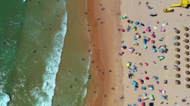Hava manzaralı ziyaretçiler, renkli şemsiyeler altında uzanarak ve dalgalarda yüzerek yaz günlerinin tadını çıkarıyorlar. Aileler ve arkadaşlar rahatlamak için toplanırken kahkaha ve eğlence ortamı dolduruyor..