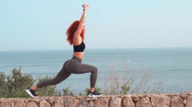 Açık gökyüzüne bakan deniz manzaralı duvarda bacaklarını açan kararlı, sportif bir kadın. Spor giysilerinde gülümseyen kadın gündüz spor yapıyor.. 
