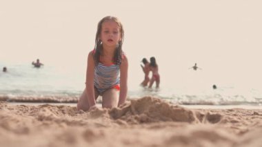 Genç kız dikkatle kumsalda kumdan kale yapmaya odaklanıyor. İnsanlar okyanusta yüzerken ve oynarken yaz güneşini emerek neşeli bir atmosfer yaratıyor..