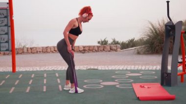 Açık spor salonunda elastik direnç bandını çeken formda bir kadın. Spor yaparken kulaklık takan aktif giysili gülümseyen genç bir kadın.. 
