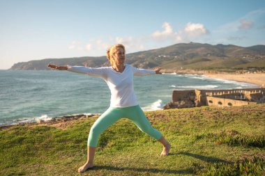 Son sınıf öğrencisi, yoga yapıyor, okyanus kıyısında asanas gösterisi yapıyor. Aktif emekli kadın vücuduna bakıyor ve güneşli bir sabahta hayatın tadını çıkarıyor. Emeklilik, rahatlama, tatil, sağlıklı yaşlı yaşam tarzı