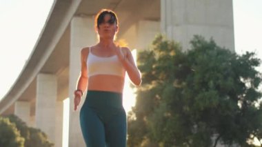 Şehir parkında güneşli bir günde köprünün altında koşan sportif kadın koşucudan. Sırtı yanan kadın koşucu spor kıyafetleriyle dışarıda egzersiz yapıyor. 