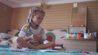 Şirin kız evinde yatağında oturup kitabı boyar gibi mutlu bir şekilde sohbet ediyor. İfadesi ve odak noktası, yaratıcılığın ve çocukluk sevincinin samimi ve iç açıcı sahnesini yaratıyor. 