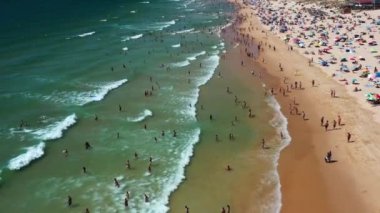 Aileler ve arkadaşlar kumlu sahilde renkli şemsiyelerle toplanırken, diğerleri ferahlatıcı okyanus dalgalarında yüzerek plaj havası manzarasında canlı bir yaz ortamı yaratıyor..