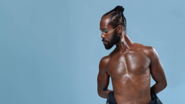 Elleri arkada mavi arka plana bakan Afro-Amerikan tişörtsüz eşcinsel model. Sade stüdyoda duran ciddi homoseksüel adam.. 