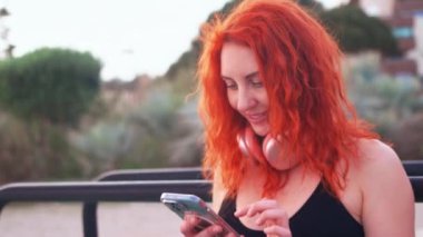 Spor giyinen kızıl saçlı kadın spor salonunda otururken akıllı telefon kullanıyor. Formda bir kadının gündüz antrenmanından sonra dinlenme görüntüsünü büyüt.