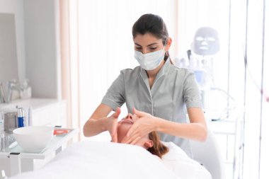 Güzellik salonunda kozmetik uzmanı kadınlara masaj yapıyor. Rahatlamış bayan müşteri güzellik uzmanları tarafından gün içinde kaplıcada şımartılıyor. Kadın profesyonel çalışırken cerrahi maske takıyor.. 