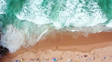 İnsanlar plaj manzarasında güneşli bir günün tadını çıkarıyorlar. Renkli şemsiyeler kumlu kıyı şeridini işaretlerken dalgalar hafifçe çarpıyor, kahkaha ve gevşemeyle dolu canlı bir yaz atmosferi yaratıyor..