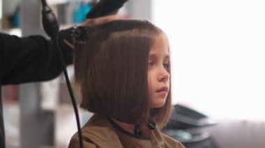 Çok tatlı bir kız berber dükkanında saçını saç kurutma makinesi ve kadın kuaförüne taratıyor. Kadın çocuk anonim berberden yeni saç stili aldı