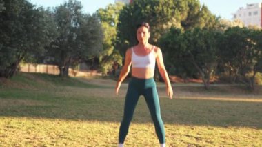 Güneşli bir günde şehir parkında zıplama pratiği yapan sportif bir kadın. Spor kıyafetli genç bir kadın gündüz vakti çimlerde egzersiz yapıyor.