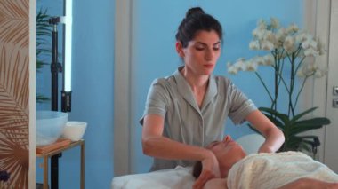 Masaj terapisti kozmetoloji kliniğinde kadınlara yüz masajı yapıyor. Profesyonel güzellik uzmanları tarafından kaplıcada şımartılan rahat bayan müşteri.