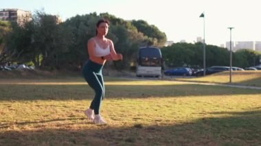 Kararlı sportif kadın güneşli bir günde çimlere atlıyor. Şehir parkında spor kıyafetleriyle ısınan sağlıklı genç bir kadın vücudu. 