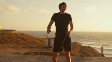 Aktif sporcu, günbatımında deniz kenarında elastik iplerle antrenman yaparken enerji dolu bir şekilde zıplar. Güç ve canlılık anı yakalar. 