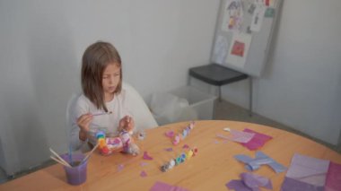 Çocukların yaratıcılık merkezinde, resim fırçaları ve yapıştırıcı kullanarak sanat ve el sanatları aktivitelerini süsleyen heykelcikleri kullanarak masada oturan bir kızın yüksek açılı görüntüsü