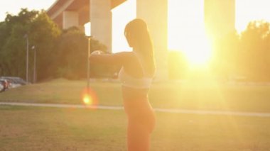 Parlak güneşli bir günde şehir parkında esneme egzersizleri yapmaya kararlı bir kadın. Arka ışıklandırmada spor giyim bölümünde genç ve formda bir kadın çimlerde egzersiz yapıyor. 