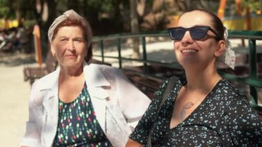Güneşli yaz parkında yaşlı annesiyle bankta oturan gülümseyen kadın, sohbetin tadını çıkarıyor, kahkaha ve anılarını paylaşıyor, birlikte geçirdikleri zamanın tadını çıkarıyor.