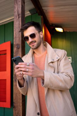Yeşil evin dışında akıllı telefon kullanarak bej renkli trençkotlu gülümseyen adam. Yakışıklı genç erkek gün boyunca tahta direğe yaslanıyor.. 