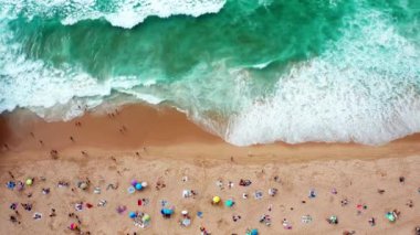 Hava perspektifi, ailelerin ve arkadaşlarının şemsiyeler altında dinlendiği hareketli bir plajı yakalar. Turkuaz dalgalar kumlu sahile çarparak güzel bir yaz günü yaratıyor..