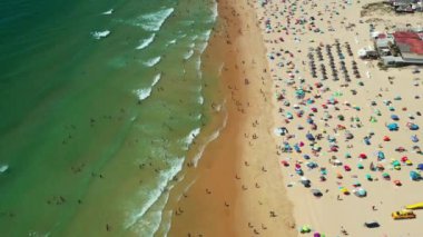 Canlı şemsiyeler altında yüzücüler ve güneş banyosu yapanlarla dolu bir kumsal. Aileler sıcak yaz atmosferinin tadını çıkarırken dalgalar yavaşça kıyıya vurur..