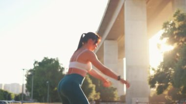 Sporty woman doing squats at city park. Determined young female athlete in activewear exercising near bridge during sunrise, focusing on strength, fitness, and healthy lifestyle