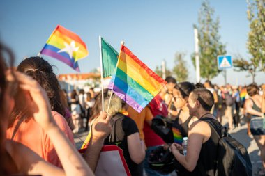 Eşcinsel erkek ve kadın kalabalığı ellerinde gökkuşağı bayraklarıyla sokaktaki gay onur yürüyüşüne katılıyor. Erkek ve dişiler güneşli bir günde açık mavi gökyüzüne karşı LGBTQ haklarını kutluyorlar. 