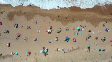 Portekiz 'in Sintra kentindeki popüler yaz mevsiminin kıyılarına deniz dalgalarının çarptığı Magoito Sahili' ndeki kumlu sahillerde insanların ve renkli şemsiyelerin insansız hava aracı görüntüsü.