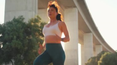 Spor giyimli kararlı bir kadın köprü altında koşuyor. Aktif genç bayan sporcu gün doğumunda şehirde egzersiz yapıyor, fitness, dayanıklılık ve sağlıklı yaşam tarzına odaklanıyor.