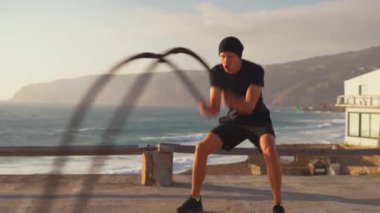 Atletik adam gün batımında deniz kenarındaki savaş halatlarıyla antrenman yapıyor. Spor, spor ve sağlıklı yaşam tarzı konsepti. Kaslı vücutlu beyaz sporcu fonksiyonel eğitim ve dayanıklılığa odaklandı
