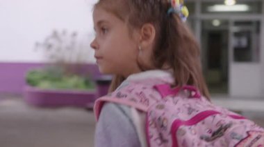 Okul dışında sırt çantalı kız öğrenci, okuldan sonra eve gidiyor, okul binası ve açık hava alanlarıyla çevrili.