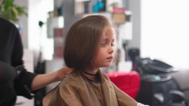 Çok tatlı bir kız berber dükkanında saç kurutma makinesiyle şekillendiriliyor ve kadın kuaför tarafından taranıyor. Anonim berberden yeni saç stili alan kadın çocuk, bu deneyimin tadını çıkarıyor.