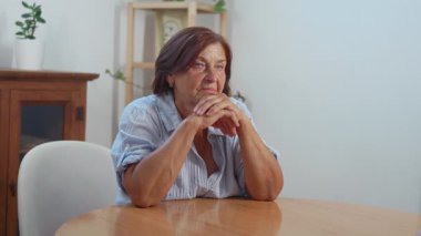 Düşünceli son sınıf öğrencisi kadın ahşap masada otururken torun şakacı bir şekilde gözlerini kapatıyor, onu kucaklıyor ve öpüyor, evde birlikte vakit geçiriyorlar.
