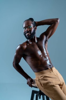 Mutlu, üstsüz siyah eşcinsel model, mavi arka plana yaslanıyor, kendine güven ve neşe sergiliyor, sıcak bir gülümsemeyle, pozitiflik ve kendini ifade etme hissi yayıyor. 