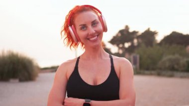 Kablosuz kulaklık takan, gülen kızıl saçlı kadın dışarıda spor salonunda müzik dinlerken eğleniyor. Egzersiz sırasında kameraya bakan mutlu atletik kadın.