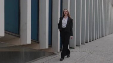 Siyah takım elbiseli genç bir iş kadını şehirdeki modern ofis binasında yürüyor. Kadın yöneticinin kendine güveni ve odaklanmış ifadesi var