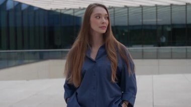 Modern ofis binasının önünde elleri ceplerinde duran koyu mavi bluzlu bir iş kadını portresi. Kameraya kendinden emin bir ifadeyle bakıyor.