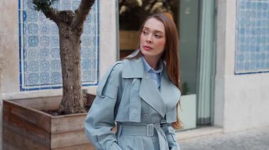 Gülümseyen iş kadını, şehir ortamında kendine güven ve sıcaklık saçan şık mavi bir trençkot giymiş kaldırım taşı caddesinde dururken neşeyle el sallıyor.