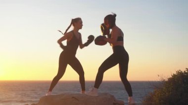 Tepede boks antrenmanı yapan sportif kadın boksör. Gün batımında deniz manzarası ve açık gökyüzü. Kadın koç ona rehberlik ediyor. Plajda kararlı boksör eğitimi.