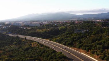 Otoyoldaki araçların hava aracı görüntüsü yemyeşil arazide dönüyor, altyapı ve doğa arasındaki uyumu gösteriyor.