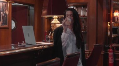 Resmi kıyafetli bir iş kadını, şık bir barda ahşap mobilya, dizüstü bilgisayar, sıcak aydınlatma ve rahat bir iş molası öneren şık bir ortamda dururken viski içiyor.