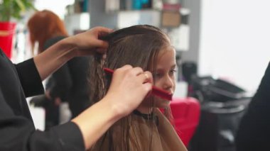 Kuaför saçlarını tararken küçük kız kuaförde saçını kestiriyor. Kuaför çocuk saçı üzerinde dikkatlice çalışıyor, düzgün ve cilalı görünmesini sağlıyor.