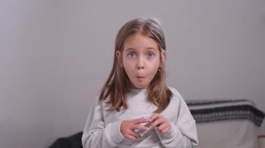 Evde dikilen, sıradan bir kazak giyen, parlak bir odada neşeli bir ifadeyle ileriye bakan, çocukluk masumiyetini, mizahını ve merakını gösteren, komik bir yüz ifadesi takınan genç kız.