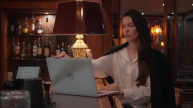 İş kadını dizüstü bilgisayar açıyor ve şık bir otel barında çalışmaya başlıyor. Odaklanmış ifade, sıcak aydınlatma, zarif ortam modern uzak çalışma ve profesyonel yaşam tarzını vurguluyor
