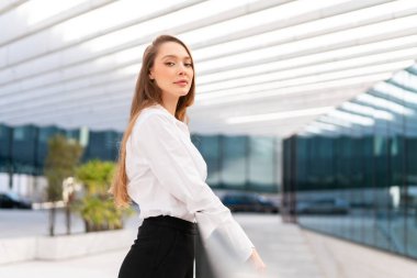 Kendine güvenen iş kadınının kameraya bakışının yan görüntüsü. Formalite içinde güzel bir kadın iş yerinde duruyor, profesyonellik yayıyor ve doğal ışıklandırmayla kurumsal ortamda duruşunu koruyor.